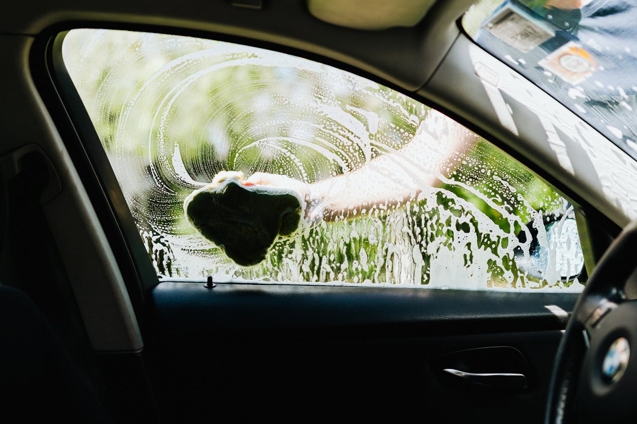 Drivers side car window being washed with a soapy sponge outdoors, capturing the cleaning process.